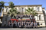 A large group of uniformed individuals is posing in front of a red fire truck with 'BOMBEIROS' written on it. They are standing and sitting in formation in front of a historic building, with palm trees adding to the scenery.
