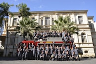 A large group of uniformed individuals is posing in front of a red fire truck with 'BOMBEIROS' written on it. They are standing and sitting in formation in front of a historic building, with palm trees adding to the scenery.