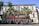 A large group of uniformed individuals is posing in front of a red fire truck with 'BOMBEIROS' written on it. They are standing and sitting in formation in front of a historic building, with palm trees adding to the scenery.
