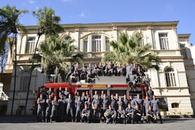 A large group of uniformed individuals is posing in front of a red fire truck with 'BOMBEIROS' written on it. They are standing and sitting in formation in front of a historic building, with palm trees adding to the scenery.