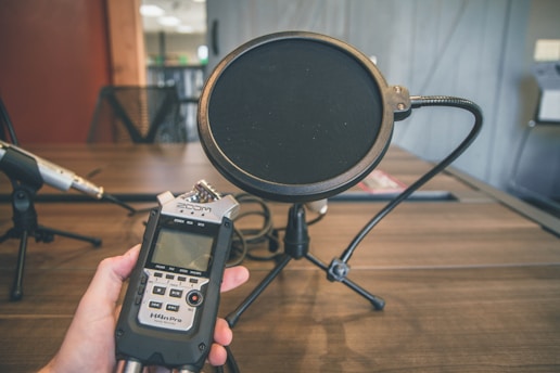 A podcast or recording setup on a wooden desk, featuring a pop filter mounted on a microphone stand and a hand-held recording device labeled H4n Pro. The workspace appears organized, with additional microphones visible in the background. The overall setting suggests an environment prepared for audio recording or broadcasting.