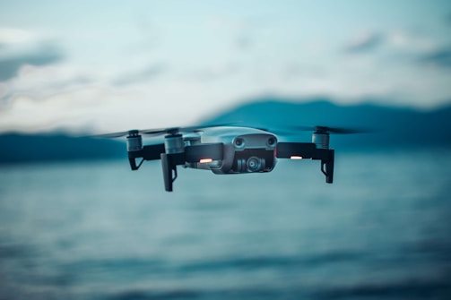 A drone hovers in the air against a blurred background of a large body of water and distant mountains. The focus is sharp on the drone's body and propellers, emphasizing its modern design and technological features. The scene is captured in natural lighting during what appears to be early morning or late afternoon, with a slightly cool tone.