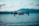 A drone hovers in the air against a blurred background of a large body of water and distant mountains. The focus is sharp on the drone's body and propellers, emphasizing its modern design and technological features. The scene is captured in natural lighting during what appears to be early morning or late afternoon, with a slightly cool tone.