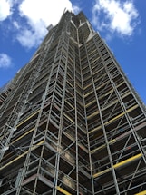 A tall structure covered in industrial scaffolding extends upward into the sky. The scaffolding consists of numerous metal bars and planks forming an intricate grid pattern. The background features a deep blue sky dotted with several white fluffy clouds.