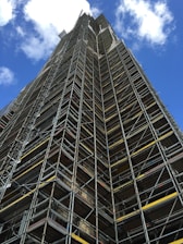 A tall structure covered in industrial scaffolding extends upward into the sky. The scaffolding consists of numerous metal bars and planks forming an intricate grid pattern. The background features a deep blue sky dotted with several white fluffy clouds.