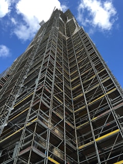 A tall structure covered in industrial scaffolding extends upward into the sky. The scaffolding consists of numerous metal bars and planks forming an intricate grid pattern. The background features a deep blue sky dotted with several white fluffy clouds.