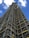 A tall structure covered in industrial scaffolding extends upward into the sky. The scaffolding consists of numerous metal bars and planks forming an intricate grid pattern. The background features a deep blue sky dotted with several white fluffy clouds.