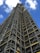 A tall structure covered in industrial scaffolding extends upward into the sky. The scaffolding consists of numerous metal bars and planks forming an intricate grid pattern. The background features a deep blue sky dotted with several white fluffy clouds.