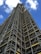 A tall structure covered in industrial scaffolding extends upward into the sky. The scaffolding consists of numerous metal bars and planks forming an intricate grid pattern. The background features a deep blue sky dotted with several white fluffy clouds.