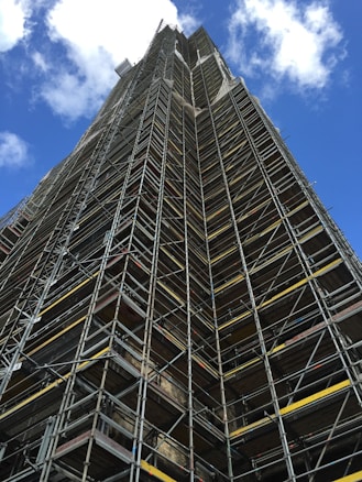 A tall structure covered in industrial scaffolding extends upward into the sky. The scaffolding consists of numerous metal bars and planks forming an intricate grid pattern. The background features a deep blue sky dotted with several white fluffy clouds.
