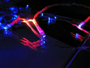 A string of decorative LED lights emitting red and blue light. The lights are intertwined, creating a vibrant and dynamic visual effect against a dark background. The glow of the lights reflects off the nearby surfaces, enhancing the vivid colors.