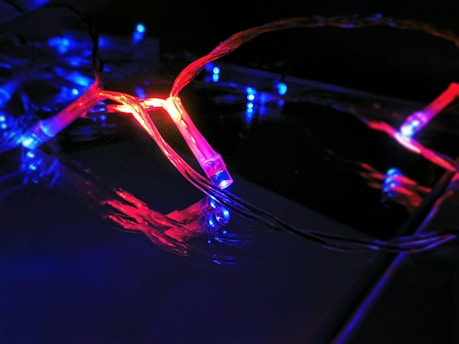 A string of decorative LED lights emitting red and blue light. The lights are intertwined, creating a vibrant and dynamic visual effect against a dark background. The glow of the lights reflects off the nearby surfaces, enhancing the vivid colors.