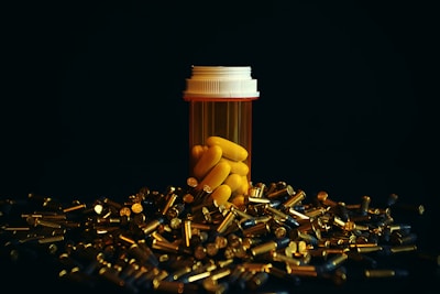 A prescription pill bottle filled with yellow tablets stands upright amidst numerous scattered bullet casings. The image is set against a dark background, highlighting the contrast between the bright yellow of the pills and the metallic sheen of the casings.