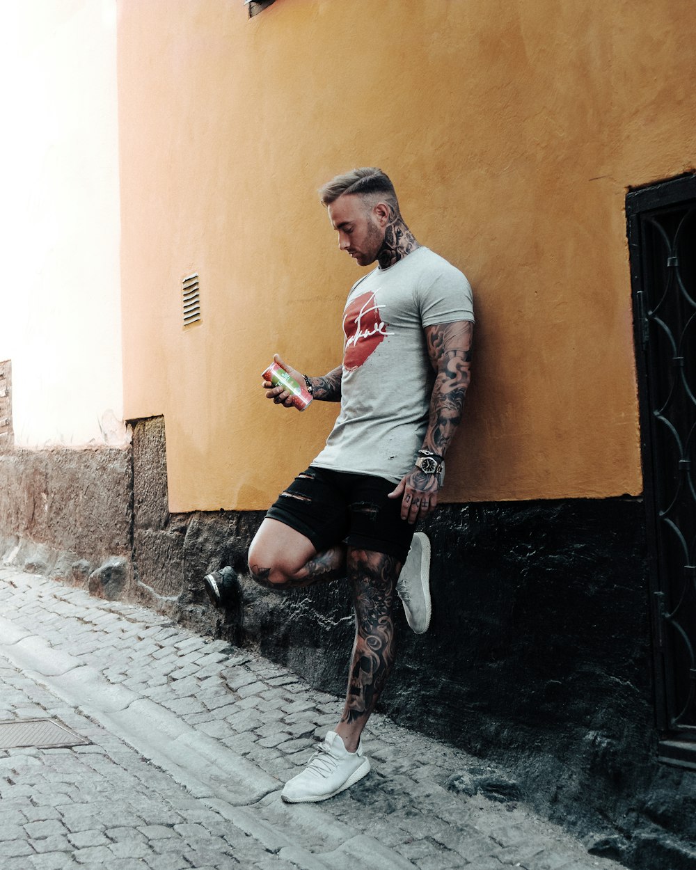 man covered with tattoo leaning on wall holding can during daytime