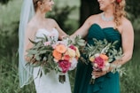 Two women outdoors, one wearing a white wedding dress with a veil and the other in a teal bridesmaid dress, both holding vibrant floral bouquets with a mix of green foliage and colorful flowers including pink, orange, and yellow roses.