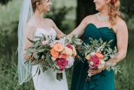 Two women outdoors, one wearing a white wedding dress with a veil and the other in a teal bridesmaid dress, both holding vibrant floral bouquets with a mix of green foliage and colorful flowers including pink, orange, and yellow roses.