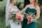 Two women outdoors, one wearing a white wedding dress with a veil and the other in a teal bridesmaid dress, both holding vibrant floral bouquets with a mix of green foliage and colorful flowers including pink, orange, and yellow roses.