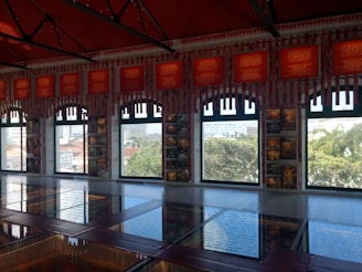 An interior space with large windows allowing abundant natural light to illuminate the room. Red-framed plaques with indecipherable text line the upper wall, while framed photographs and artwork are displayed between the windows. A reflective surface on the floor mirrors the windows and exterior view, which includes trees and distant buildings.