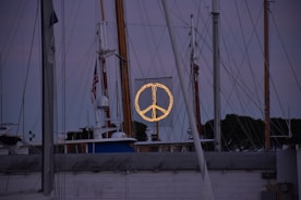 A marina scene with multiple sailboats and yacht masts set against a dusk sky. In the center, a large illuminated peace symbol is displayed on a mesh background. The surrounding area is dimly lit, creating a calm and serene atmosphere. An American flag is visible on one of the masts.