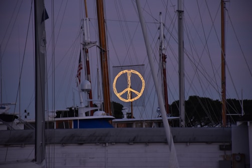 A marina scene with multiple sailboats and yacht masts set against a dusk sky. In the center, a large illuminated peace symbol is displayed on a mesh background. The surrounding area is dimly lit, creating a calm and serene atmosphere. An American flag is visible on one of the masts.
