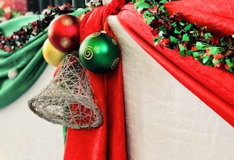 Festive decorations featuring bright red and green drapery adorned with shiny Christmas ornaments, including red, green, and yellow spheres with decorative patterns, along with a silver wire bell. A garland of mixed red and green tinsel embellishes the scene.