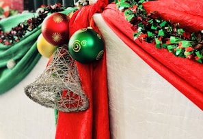 Festive decorations featuring bright red and green drapery adorned with shiny Christmas ornaments, including red, green, and yellow spheres with decorative patterns, along with a silver wire bell. A garland of mixed red and green tinsel embellishes the scene.
