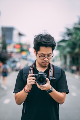 An individual wearing glasses, dressed in a black shirt, is holding a DSLR camera. A camera strap is visible around the neck. The background appears to be an urban street scene, slightly blurred, with buildings and greenery.