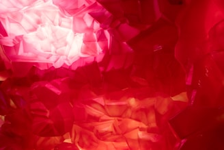 A close-up view of a vibrant, translucent red and pink crystal structure, with intricate facets that reflect and refract light, creating a glowing appearance.