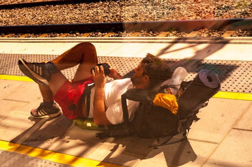 A man is lying on his back on a train platform, using a backpack as a pillow. He is wearing a white shirt, red shorts, and sneakers, and is holding a camera in his hand. The backpack is equipped with a water bottle, a rolled-up sleeping mat, and other essentials. The background shows railway tracks, with sunlight casting shadows on the platform.