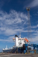 A large cargo ship is docked at a port under a bright blue sky with scattered clouds. The vessel is equipped with orange lifeboats and has white structures on its deck. A tall metal tower is positioned nearby, possibly for lighting or communications.