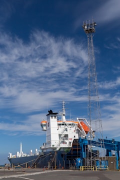 A large cargo ship is docked at a port under a bright blue sky with scattered clouds. The vessel is equipped with orange lifeboats and has white structures on its deck. A tall metal tower is positioned nearby, possibly for lighting or communications.