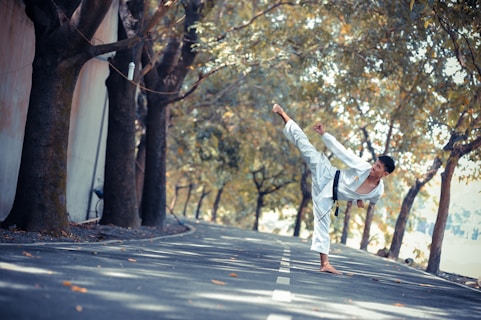 A person wearing a white martial arts uniform performs a high kick on a tree-lined road. The trees cast shadows on the asphalt, and fallen leaves are scattered around. The setting seems calm and serene, with a focus on precision and balance in the martial arts pose.