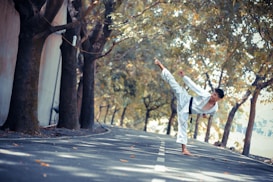 A person wearing a white martial arts uniform performs a high kick on a tree-lined road. The trees cast shadows on the asphalt, and fallen leaves are scattered around. The setting seems calm and serene, with a focus on precision and balance in the martial arts pose.