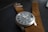 A close-up view of a wristwatch with a brown leather strap placed on a textured black surface. The watch face is dark with white numbers and has a silver metallic casing. The strap has a series of evenly spaced holes and a metallic clasp.