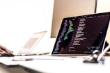 A dimly lit office setting with a laptop displaying code on the screen. The image shows a programming environment with various lines of HTML or JavaScript. A blurred hand is using a mouse on the left side, while another laptop is partially visible in the background. The overall setting suggests a workspace focused on coding or software development.