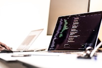 A dimly lit office setting with a laptop displaying code on the screen. The image shows a programming environment with various lines of HTML or JavaScript. A blurred hand is using a mouse on the left side, while another laptop is partially visible in the background. The overall setting suggests a workspace focused on coding or software development.