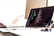 A dimly lit office setting with a laptop displaying code on the screen. The image shows a programming environment with various lines of HTML or JavaScript. A blurred hand is using a mouse on the left side, while another laptop is partially visible in the background. The overall setting suggests a workspace focused on coding or software development.