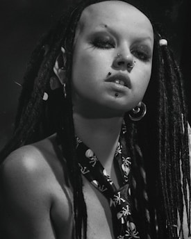 A person with long black dreadlocks and multiple facial piercings, including a nose and lip piercing. They are wearing a skull-patterned necktie and large hoop earrings. The image is in black and white, giving it a dramatic and bold look.