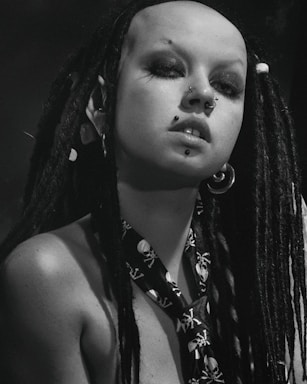 A person with long black dreadlocks and multiple facial piercings, including a nose and lip piercing. They are wearing a skull-patterned necktie and large hoop earrings. The image is in black and white, giving it a dramatic and bold look.