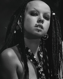 A person with long black dreadlocks and multiple facial piercings, including a nose and lip piercing. They are wearing a skull-patterned necktie and large hoop earrings. The image is in black and white, giving it a dramatic and bold look.