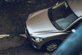 An overhead view of a metallic silver car parked on a textured, asphalt surface. The car’s windshield and hood are visible, with reflections indicating a cloudy day. The image captures part of a street line and the edge of a pedestrian walkway, creating an urban setting.