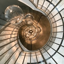 A spiral staircase with a modern design, featuring glass balustrades and metal railings. The view is looking downwards from a high vantage point. At the center, several spherical light fixtures are suspended, creating an artistic and elegant focal point.