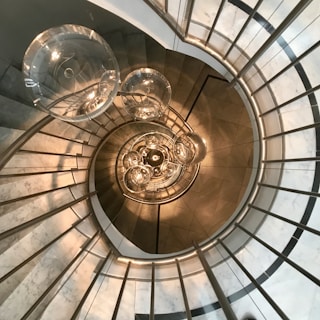 A spiral staircase with a modern design, featuring glass balustrades and metal railings. The view is looking downwards from a high vantage point. At the center, several spherical light fixtures are suspended, creating an artistic and elegant focal point.