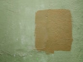 A textured wall with a light green color and a rectangular patch of a darker tan paint applied over it. The paint appears uneven, adding a rough texture to the surface.