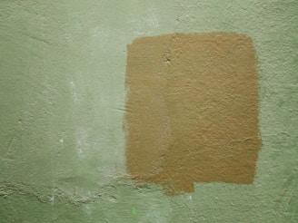 A textured wall with a light green color and a rectangular patch of a darker tan paint applied over it. The paint appears uneven, adding a rough texture to the surface.