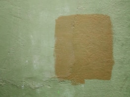 A textured wall with a light green color and a rectangular patch of a darker tan paint applied over it. The paint appears uneven, adding a rough texture to the surface.