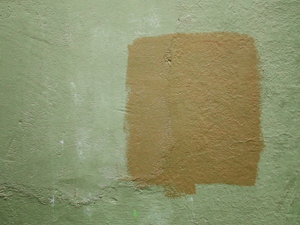A textured wall with a light green color and a rectangular patch of a darker tan paint applied over it. The paint appears uneven, adding a rough texture to the surface.
