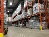 A warehouse with tall, orange metal shelving units filled with pallets wrapped in plastic. The floor is concrete, and overhead industrial lighting illuminates the space. The shelving reaches up to the ceiling, maximizing storage capacity with numerous boxes and items stacked neatly.