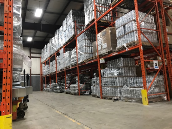 A warehouse with tall, orange metal shelving units filled with pallets wrapped in plastic. The floor is concrete, and overhead industrial lighting illuminates the space. The shelving reaches up to the ceiling, maximizing storage capacity with numerous boxes and items stacked neatly.