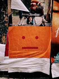 Layered posters on a wall include an advertisement featuring two people in casual, colorful clothing, with one person lying on a wooden deck. Below, an abstract poster depicts a minimalist orange face with a neutral expression, composed of simple red shapes.